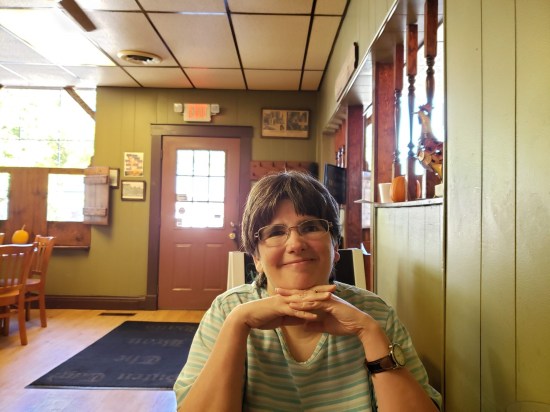 Anne smiling at me across the table inside a restaurant with green and brown decor. Sunlight pours in a window at left.