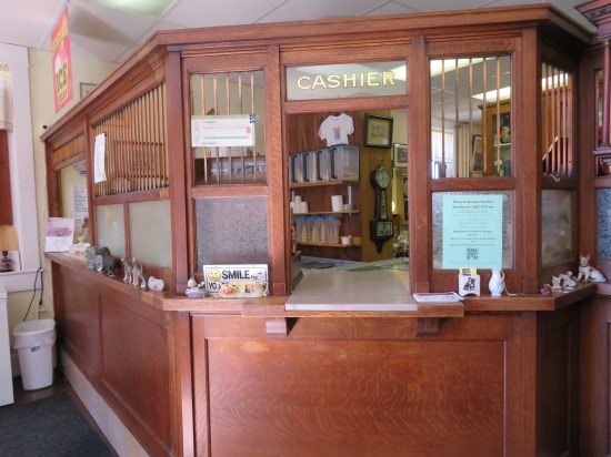 Inside is an old wooden bank teller window, complete with CASHIER window pane at the top. Non-bank objects clutter around such as shirts, clocks, and Tupperware holding ice cream cones.