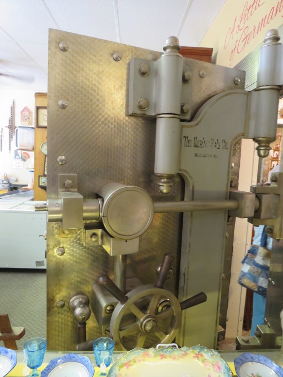 An entire, open, massive bank vault door. Old cups and plates are on a shelf in front of it.