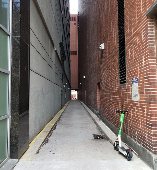 Long alley with a scooter leaning against one brick wall. At the far end, barely visible is a Frisch's Big Boy sign, possibly on a truck.