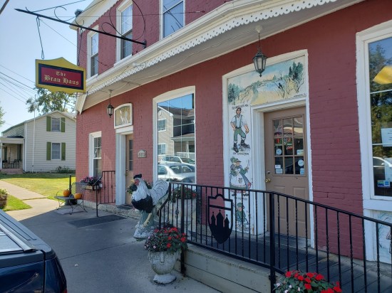 Angled view of the front of a brick restaurant with a red-and-yellow THE BRAU HAUS sign hanging on a pole bracketed on the wall. Decor includes a black iron fence with a silhouetted crest of spires, German Oktoberfest celebrants painted around the door, and a chicken statue.