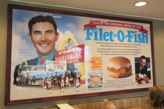 Large indoor placard detailing the history of the Filet-O-Fish tribute, with a painting of inventor Lou Groen. A customer's head is in the shot.