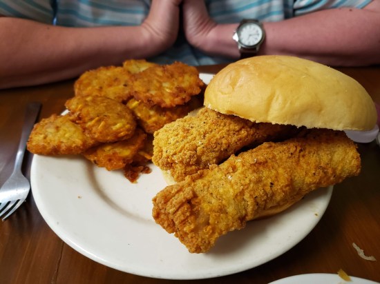 On a dinner plate is a pile of small potato pancakes, and two large fried fish fillets on a bun.