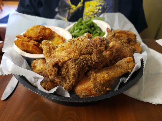 A basket filled with five pieces of fried chicken. Behind it is a plate with green beans and potato pancakes.