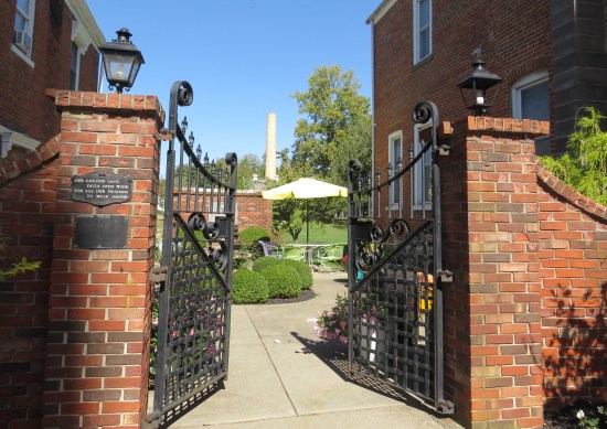 A brick wall with a metal gate opened up to a patio with bushes, table, patio umbrella, chairs and trees, In the distance is a lone, tall factory smokestack jutting into the horizon.