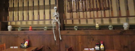 A wooden shelf above two restaurant tables with a skeleton and several small skulls lined across it.