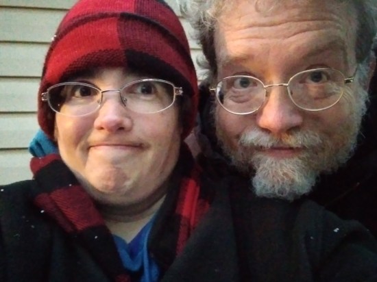 Selfie! My head looking directly into the camera and smiling. Next to me, Anne is making a frustrated face at her uncooperative camera while side-eying me. She's wearing matching red flannel cap and scarf. She cut the top of my head out of the shot.