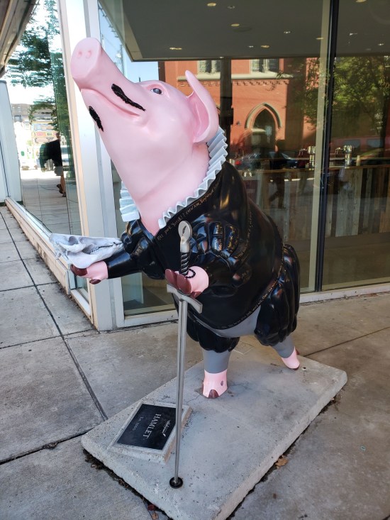 Statue of a pig in a black Elizabethan suit with Shakespeare quotes written on it. The pig has a thin mustache and props itself up on a sword.
