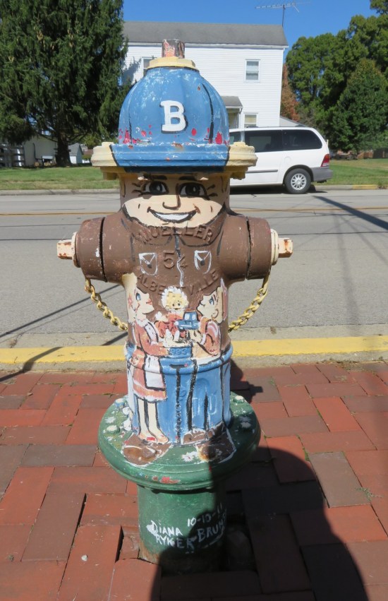 Fire hydrant painted like a man in brown factory wear and a blue hat with a 'B' on it, plus a family standing in front of him.