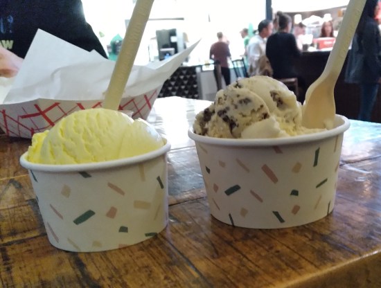 Closeup of two cups of ice cream on a wooden table. The ice cream stand and its two employees are a white, overlit blur in the background.