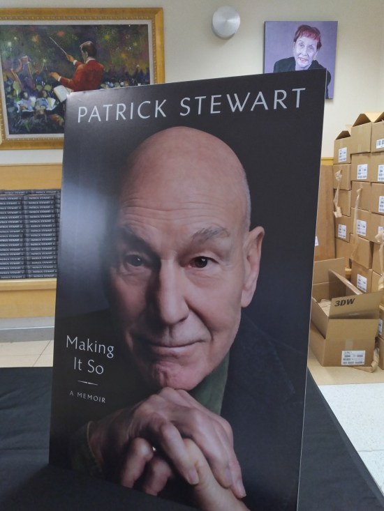 A copy of "Making It So" standing atop a pile of others. Far background, student art hangs on the white wall, including a painting of an orchestra conductor evocative of Van Gogh's "Starry Night".
