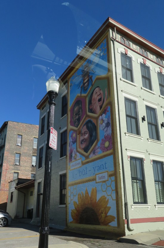 A blue vertical mural with pics of three Spelling Bee kids in beehive hexagons with the dictionary entry for the word EBULLIENT at the bottom and a big bee at the top.