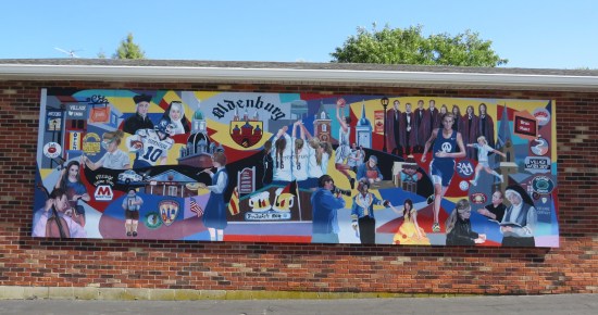 Mural on the side of a brick building with the Oldenburg crest, local business signs, and citizens from various professions (sports, science, drama, clergy, etc.).