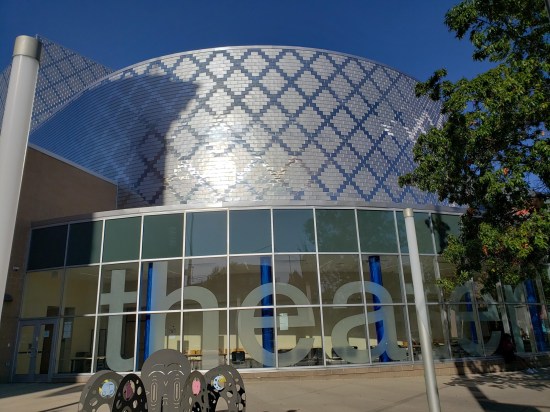 A metal cylindrical portion of a large school. The ten-foot windows have six-foot letters in lowercase: "theater".