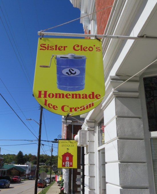 Wooden yellow sign with red lettering for Sister Cleo's Homemade Ice Cream, hung sideways from a bracket on the brick building. Center image is a drawing of a blue ice cream maker.