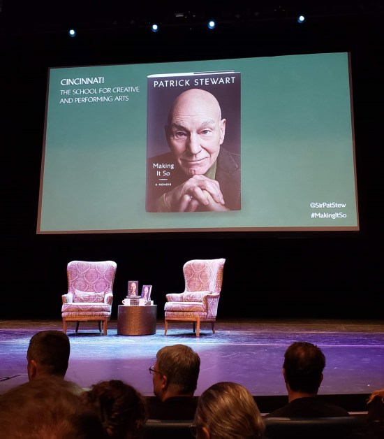 Two empty drawing room chairs on a purple-lit stage. Between them is a table with two mugs of water and two small piles of Stewart's memoir. Projected on the back wall is a photo of the book, Upper-left screen corner says "Cincinnati: The School for Creative and Performing Arts". Lower right corner has Stewart's Twitter handle @SirPatStew and the suggested hashtag #MakingItSo.