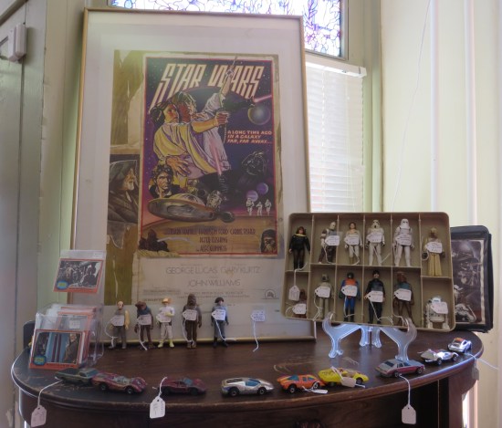 A framed old Star Wars poster by Drew Struzan and Charlie White III (possibly a reprint) stands on a shelf along with a couple dozen loose Star Wars figures and some unrelated Hot Wheels.