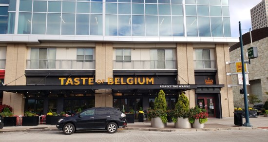 Restaurant called Taste of Belgium in the bottom floor of a tall building with glass windows reflecting the cloudy blue sky. Several large planters provide copious greenery, albeit obscured by an SUV parked out front and kinda ruining the shot.