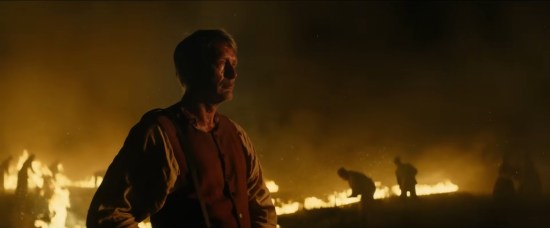 Period-piece farmer Mads Mikkelsen standing over a burning field at night.