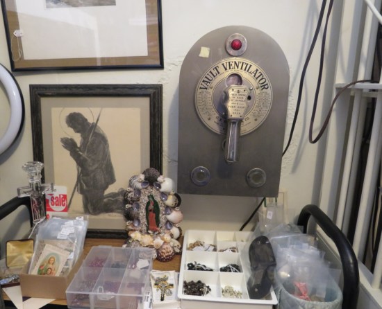 A Vault Ventilator hanging on the wall, is a metal cabinet with a red light and a handle to pull to let in air. Below it, a shelf holds Catholic jewelry and small pictures.