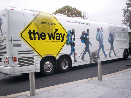 White charter bus with "The Way" movie logo on a yellow diamond road sign. Pictures of the four main cast members show them hiking toward the front of the bus.