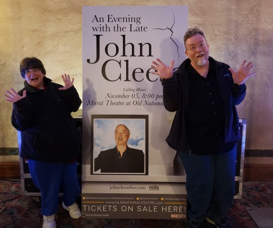 Me and Anne doing jazz hands next to a cardboard standee advertising the event, with a photo of the elderly Cleese in the center. I'm blocking the last two letters in Cleese's name. 