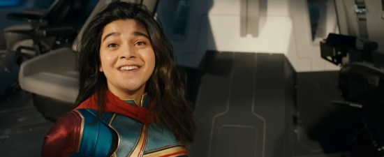 Iman Vellani in costume as Ms. Marvel, standing in a spaceship cockpit and smiling starstruck at an off-screen Captain Marvel.