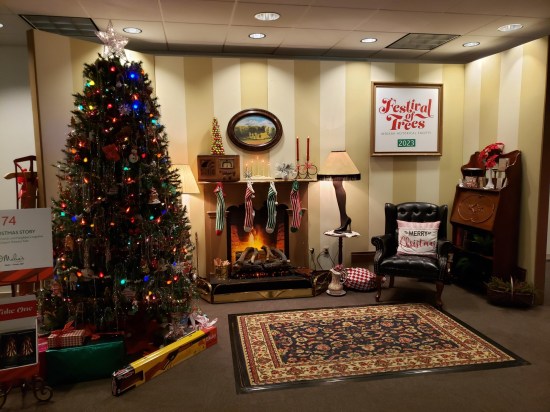 Museum exhibit of a faux living room with Christmas tree, black chair, fake fireplace, old-time radio, leg lamp, Persian rug, and a Red Ryder BB gun under the tree in its box. A "Festival of Trees 2023" wall sign is the most noticeable anachronism.
