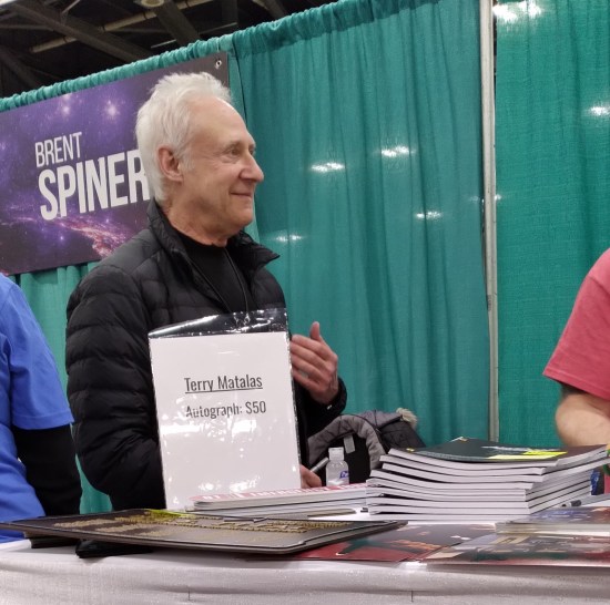 A smiling Brent Spiner stands between his table and Terry Matalas', discussing their Friday night plans.