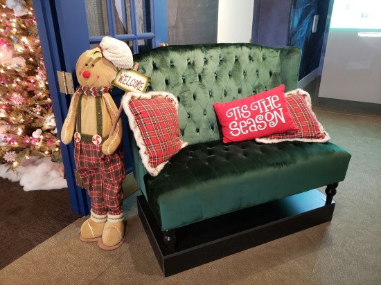 A green couch in a museum hallway with three red Christmas pillows. One of them says "'Tis the season". Next to it is a sort of gingerbread scarecrow wearing plaid Christmas pants and suspenders with peppermint buckles, holding a sign reading "Welcome".