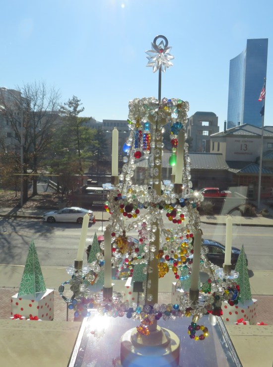 Collection of crystals hanging from a custom rack shaped like a desktop Christmas tree. A window behind it looks upon downtown Indianapolis, including a Marriott hotel shaped like a blast shield.