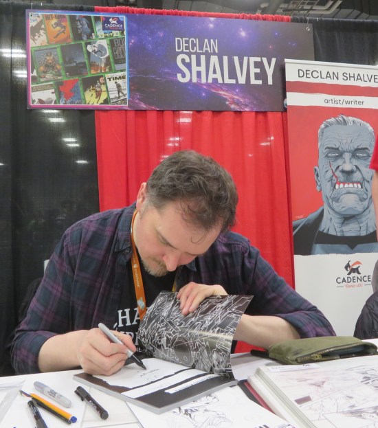 Declan Shalvey at his table, sketching in the front page of a graphic novel. His name is on a large card mounted above red curtains behind him. A standee at right promotes his Image series "Old Dog".