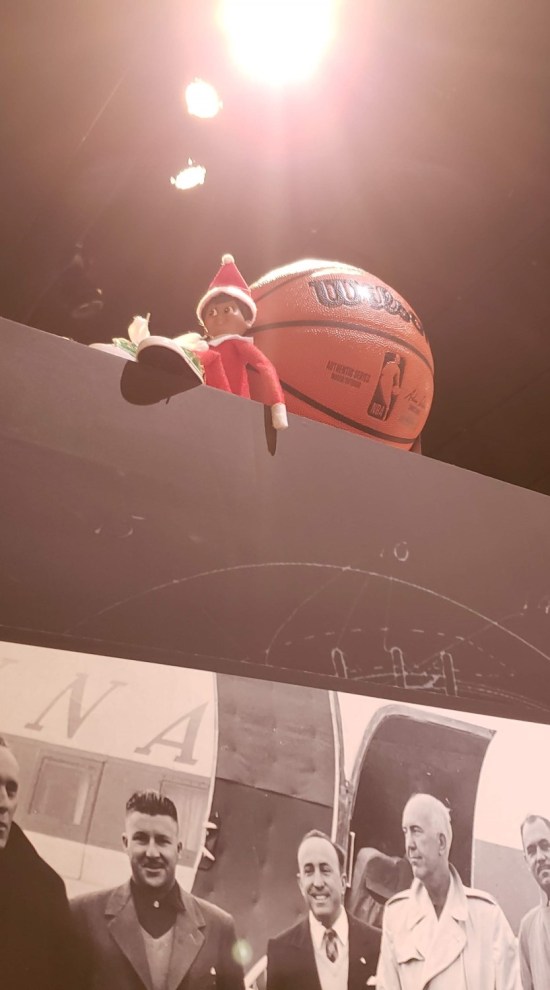 An Elf on the Shelf on a tall wall, sitting next to a basketball. Below them hangs a black-and-white photo that probably has Chuck Taylor in it.