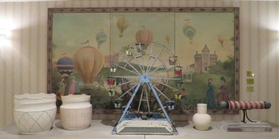 Antique Ferris wheel model on a white table in front of a painting described in the caption.