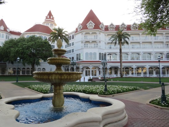 A fountain in an outdoor courtyard in front of a fancy four-story hotel with red roofs and palm trees out front.