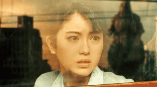 Japanese woman on a train looking out the window, which hazily reflects a rampaging Godzilla heading her way.