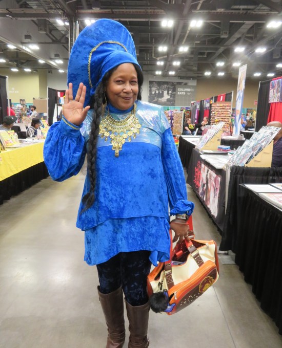 Woman in blue Guinan costume with appropriately large jewelry.