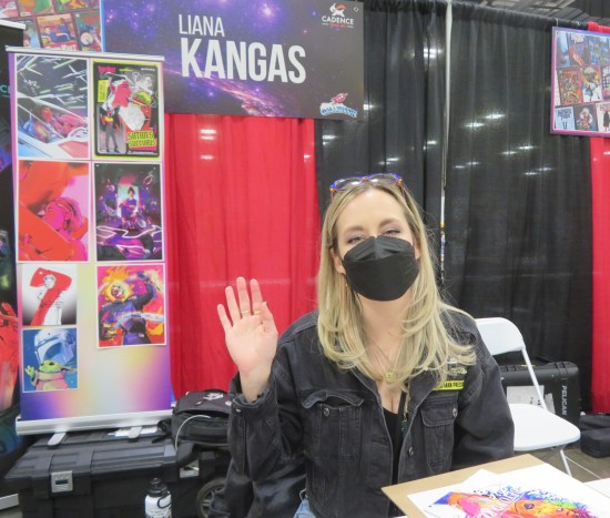 Liana Kangas at her table, waving and wearing a black mask. Art samples are on a board behind her.