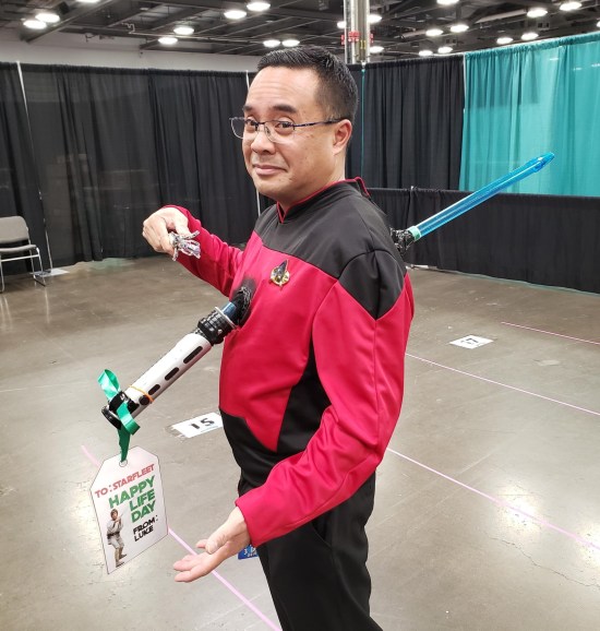 Star Trek crewman with a lightsaber through his chest. On its handle is a gift tag reading "To Starfleet, Happy Life Day, From Luke." He's also holding a broken Sonic Screwdriver.