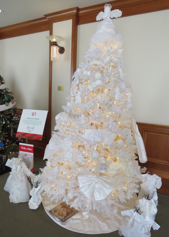 All-white Christmas tree with white ribbons, topper, and baby gowns standing around it.