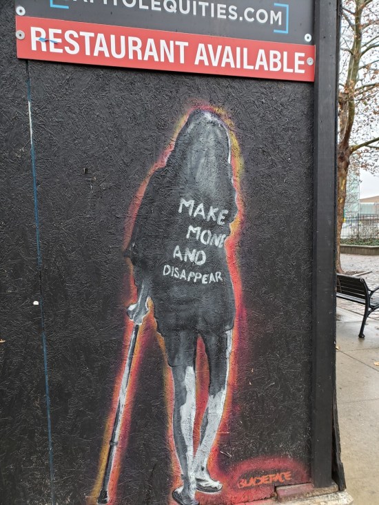 Street art: a shadowy hooded figure leans on a cane and wears a shirt saying "Make Money and Disappear". The figure is painted on black plywood on an empty storefront with a sign advertising "restaurant available".