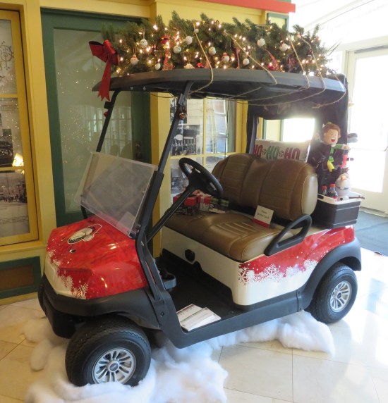 Golf cart parked indoors on fake snow stuffing, The car is red with white snowflake trim. An elf doll sits on the back. A Christmas tree is lying on the roof and tied down.