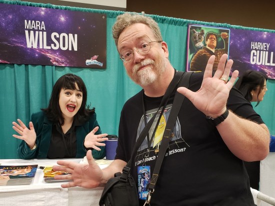 Me looming large in front of Mara Wilson's table while she remains seated behind it and very, very tiny in comparison. We're both excitedly doing jazz hands, of course. She's in a black dress and wearing makeup, including bold red lipstick.