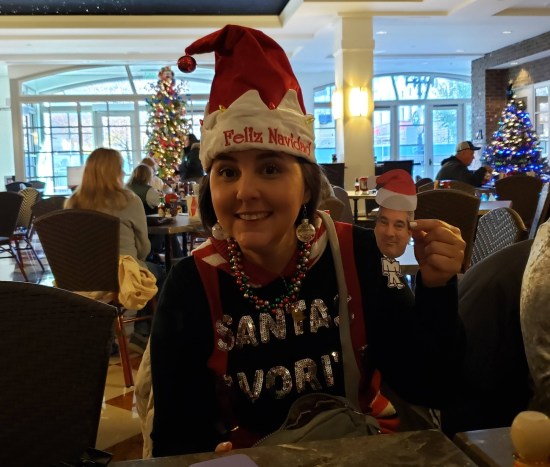 Woman in a Santa hat that says "Feliz Navidad", shirt that says "Santa's Workers", and silver ball-ornament earrings.