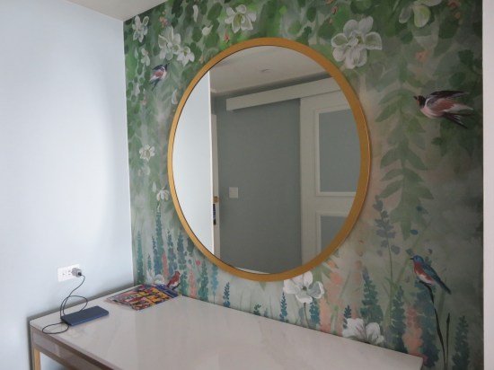 Circular mirror on a wall with green-forest wallpaper with birdies painted into it. Below is a white table with my wife's phone charging and a few comics catalogs I brought along to skim and discard.