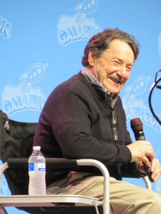 Peter Cullen on stage with a mic, seated and smiling.