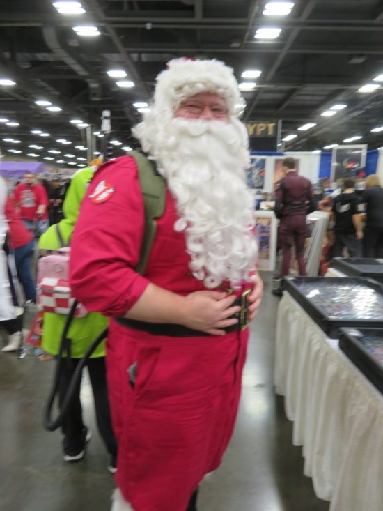 Santa in a red jumpsuit with a Ghostbusters symbol on the shoulder and a lime green proton pack.