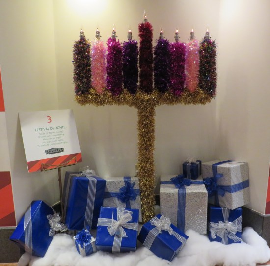 A five-foot-tall faux menorah sheathed in six kinds of garland. Around its base are presents wrapped in blue and white, sitting on a white blanket.