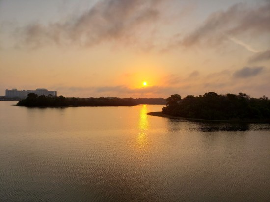 Sunrise over the lagoon, lots of orange sunlight filtered through sparse clouds. Disney's Contemporary Resort is on the far left horizon.