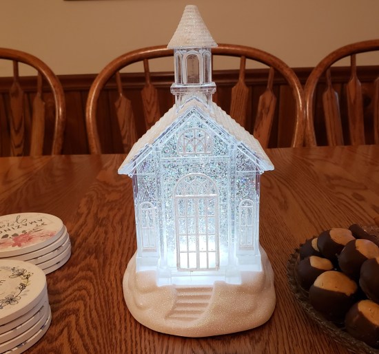 Tiny glass church with constantly moving sparkles like a snow globe. It sits on a wood dining table. Nearby are buckeye fudge balls and Christian-themed drink coasters.